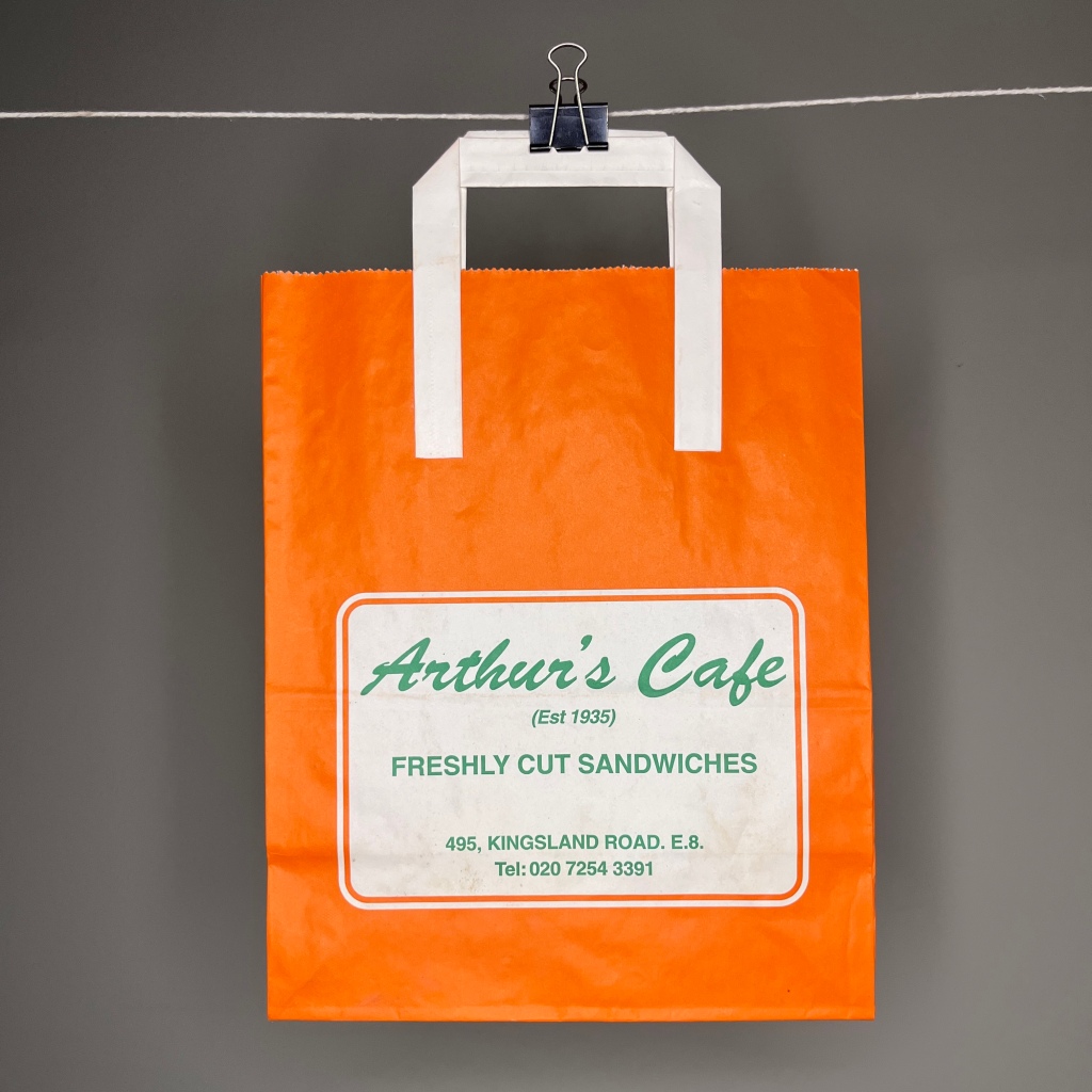 Hanging off a white string by a bulldog clip. A bright orange paper bag with paper handle. In greed curly script the bag says Arthur's Cafe and under in all caps Est 1935 Fresh Cut Sandwiches 495 Kingsland Road E8 02072543391