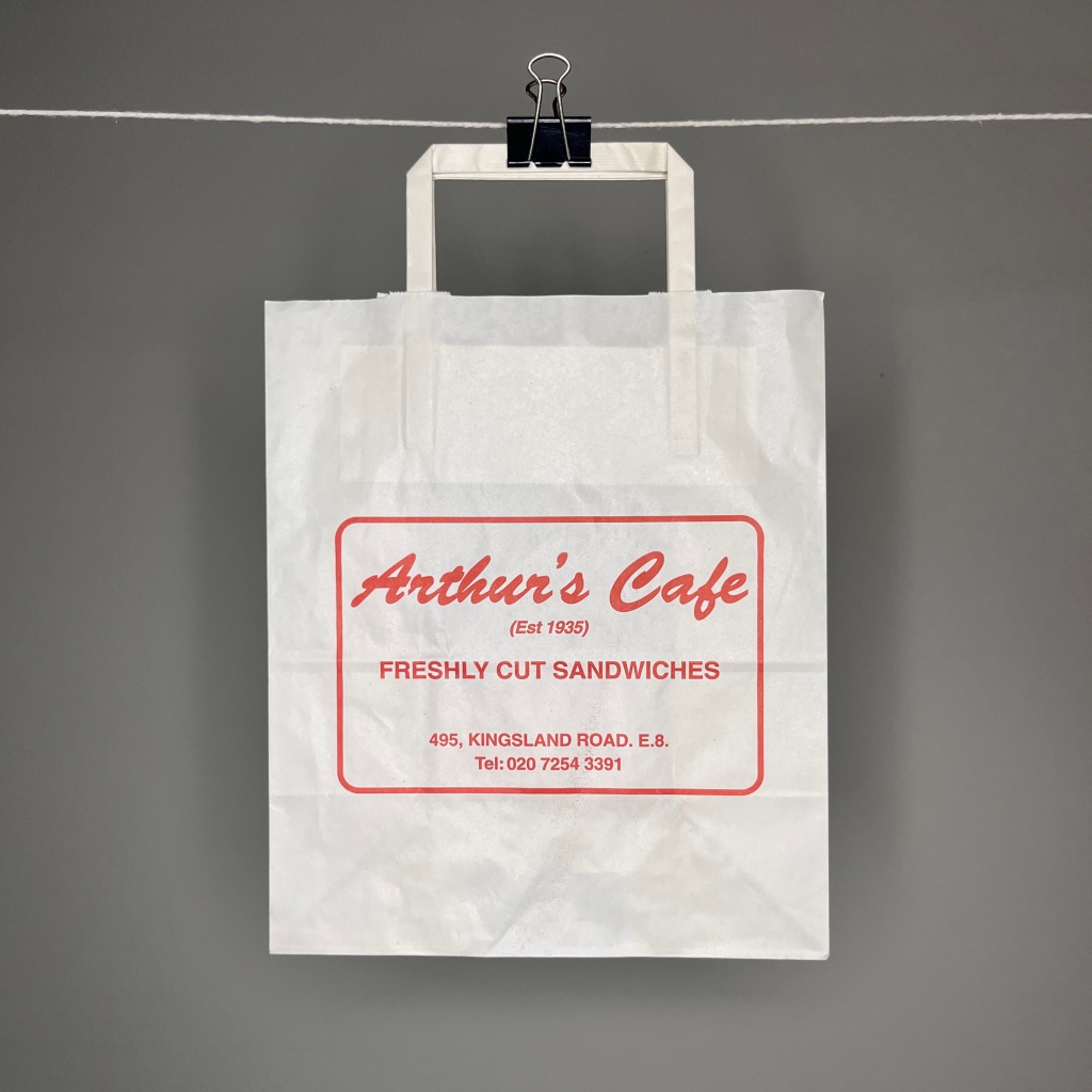 Hanging off a white string by a bulldog clip. A white paper bag with paper handle. In red curly script the bag says Arthur's Cafe and under in all caps Est 1935 Fresh Cut Sandwiches 495 Kingsland Road E8 02072543391