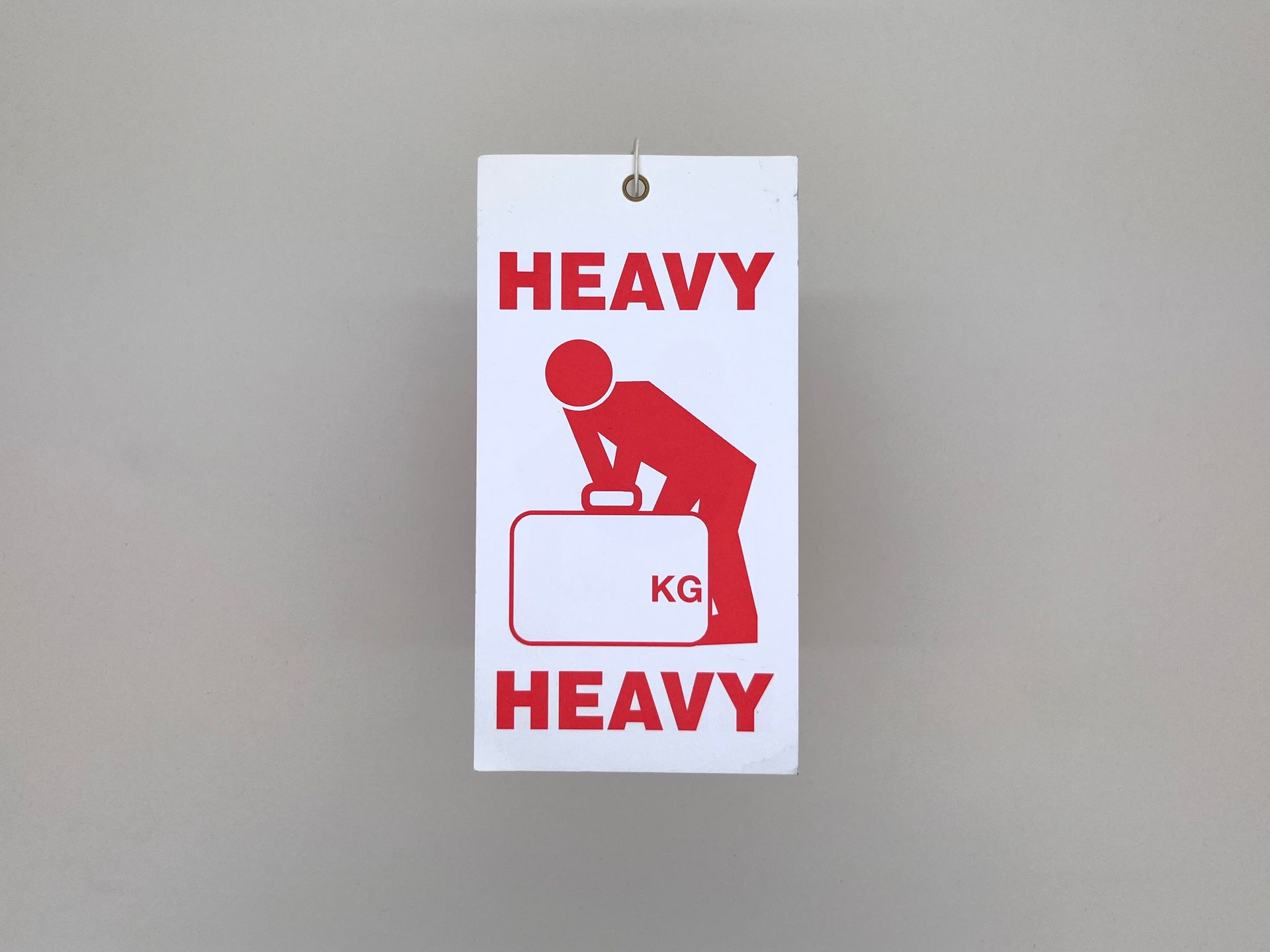 A white luggage label that says HEAVY in red capital letters above and below a red icon of a person struggling to lift a suitcase with KG written on it.