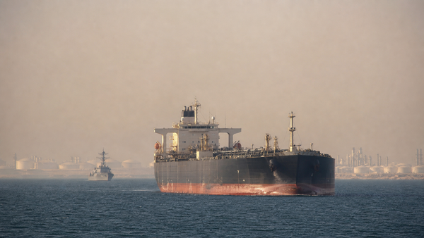 A crude-oil tanker crosses the Strait of Hormuz with a naval escort and refinery infrastructure in the distance.
