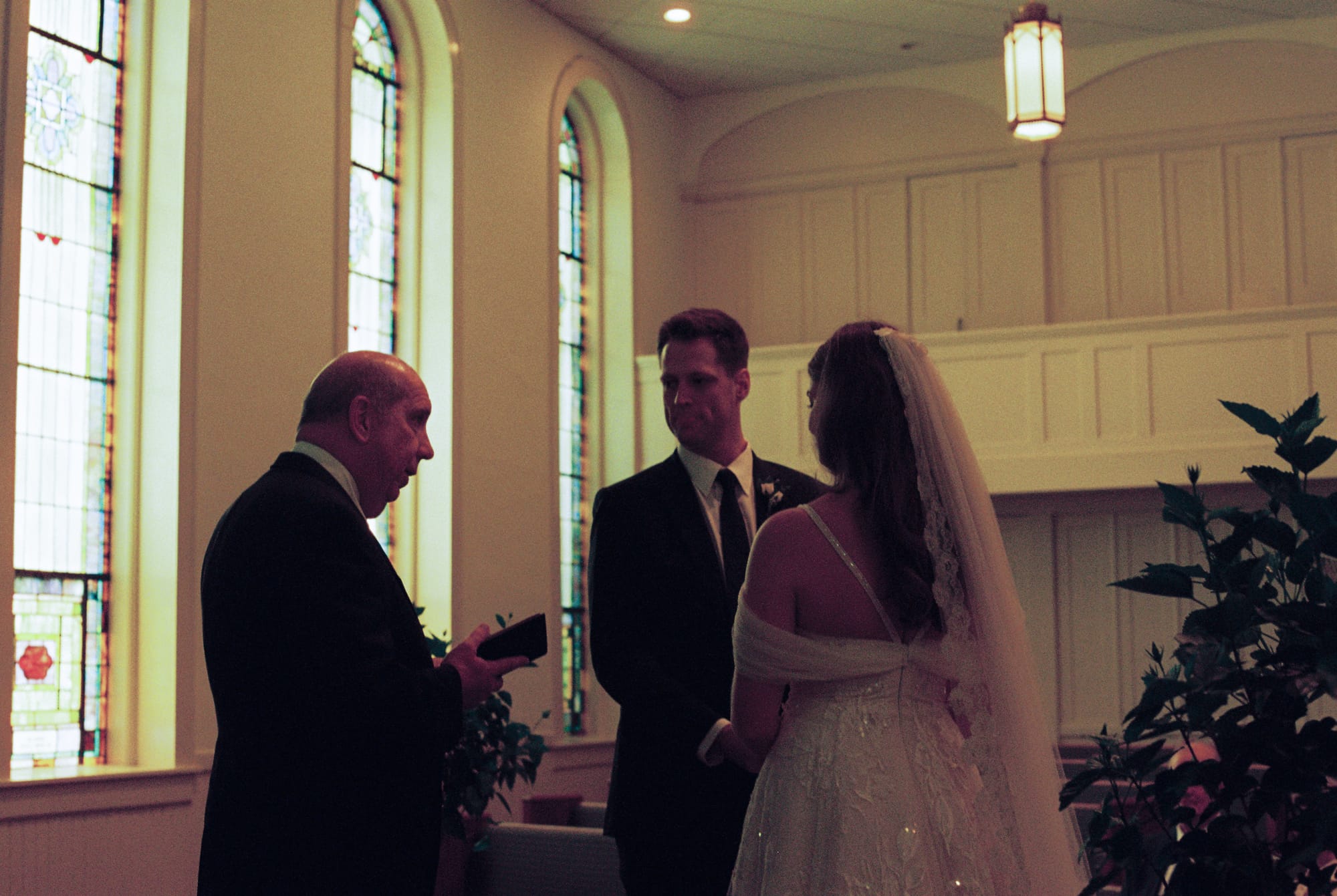 A Bride and groom inside of a nearly empty church as the pastor officiates their wedding ceremony. Tall stained glass windows line the walls in the background. 
