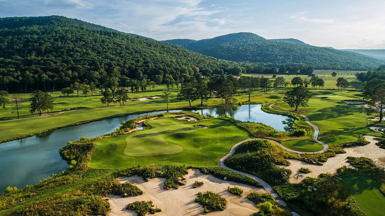 Aerial view of a green bordered by water at Sweetens Cove Golf Club in Tennessee, with sandy waste, native grass, a curving cart path, and forested ridges.