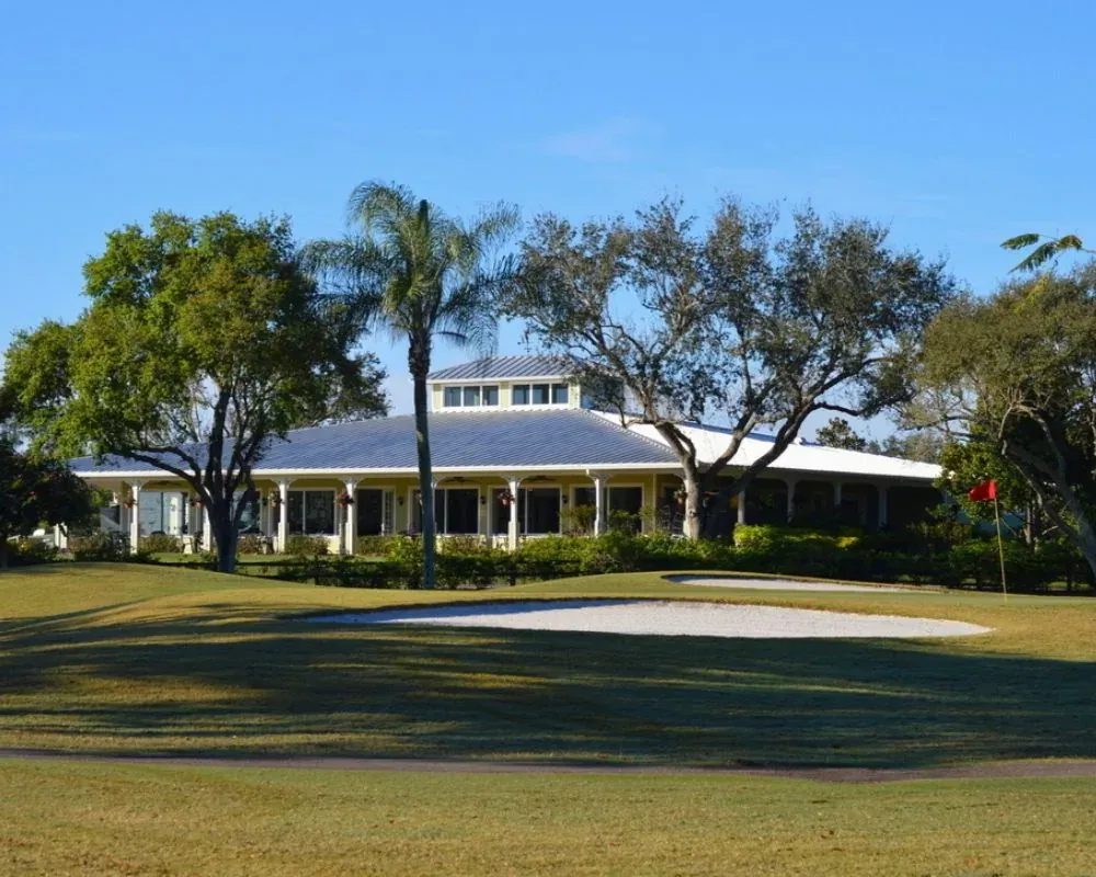 The Village Golf Club clubhouse, a low building with a metal roof and wide porch, seen across a bunker and green with a red flag