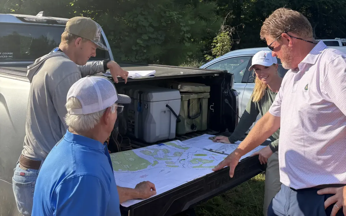 Architect Kris Spence and members of the High Side team study a course plan spread across a pickup truck tailgate at the Kannapolis site, with woods behind