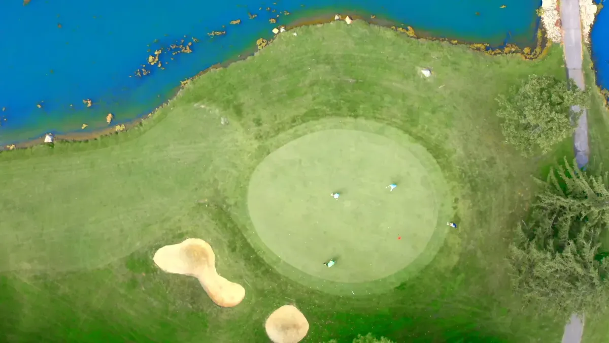 Aerial view looking straight down on a round green with golfers putting, two sand bunkers, and a pond along the fairway at Marion Country Club