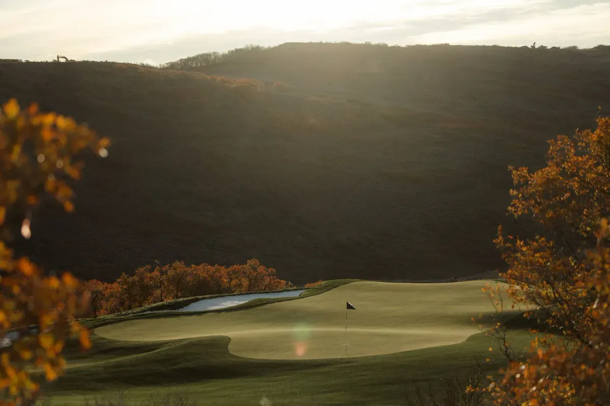 Backlit late-day view across a putting green with a black flag at Marcella Club in Utah, pond at left, orange autumn trees both sides, brush hillside, pale sky.
