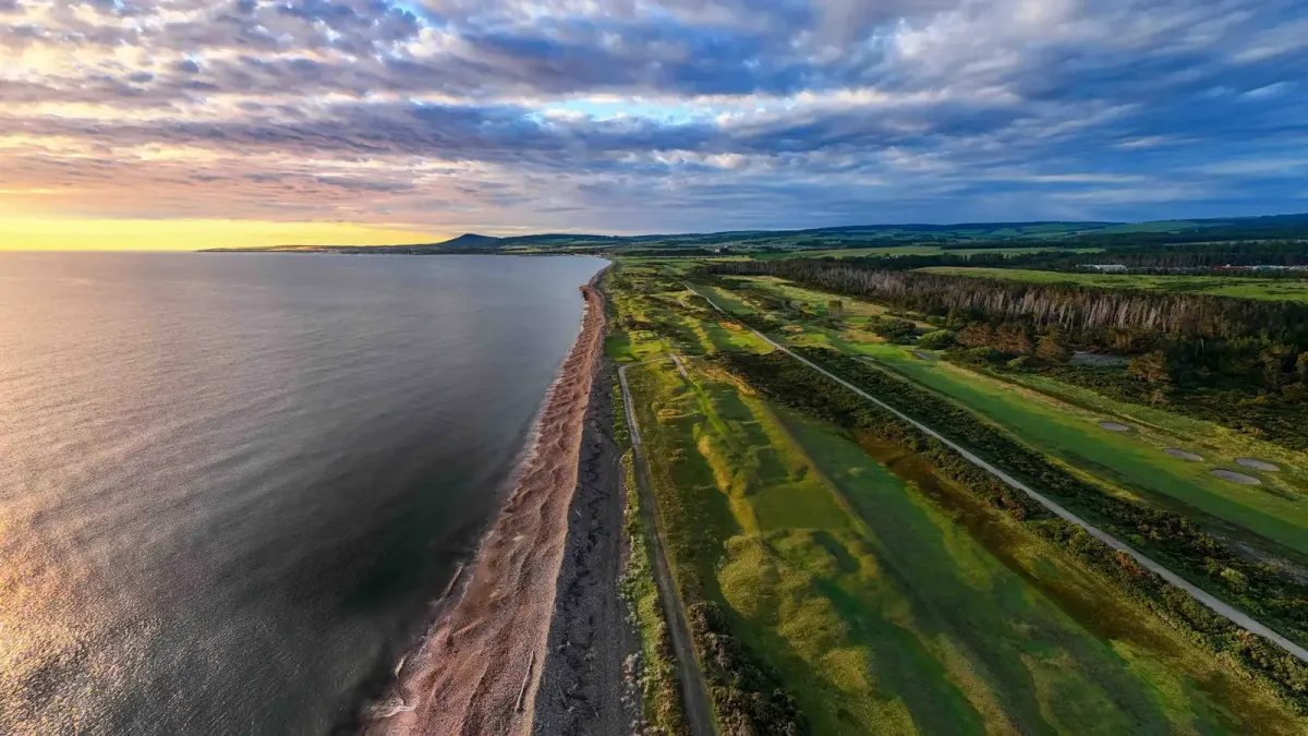 Aerial view of Spey Bay Golf Club links along the Moray Firth coast in Scotland at low sun, fairways beside the shoreline with hills inland.