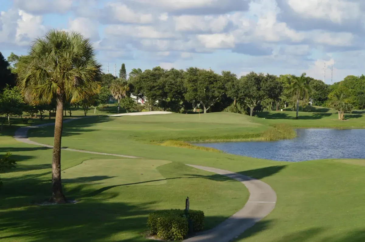 A palm tree and cart path frame the waterside 14th hole at Village Golf Club, with a pond, a green, and oak trees under a cloudy sky