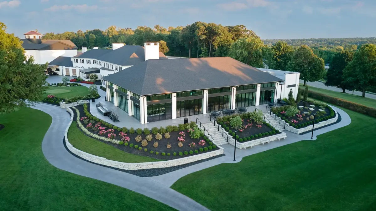 Aerial view of the event pavilion at Westbrook Country Club, with tall glass walls, a stone terrace, planted beds, and the existing clubhouse behind it