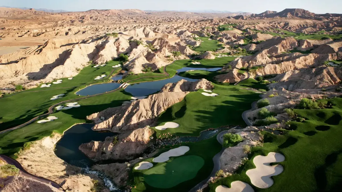 Aerial of Wolf Creek Golf Club: green fairways, white bunkers, and blue ponds between eroded tan badland ridges and canyons under a pale sky.