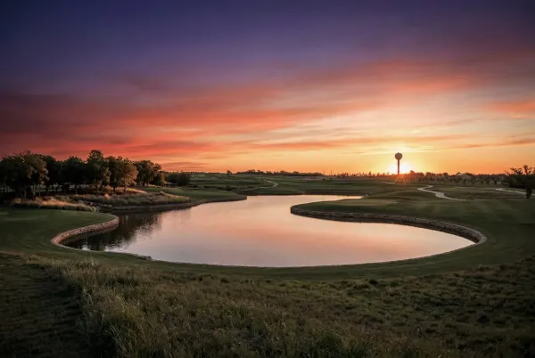 A lake ringed by a low stone wall reflects a sunset sky at Bridgesong, with fairways, a cart path, and a water tower on the horizon