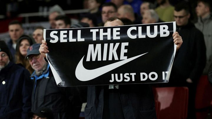football fan holding up a sign that reads "sell the club Mike" with the Nike logo and "just do it"