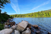 Rocky shoreline along Jordan Pond in Acadia National Park, with clear blue water, dense evergreen trees, and mountain slopes in the background.