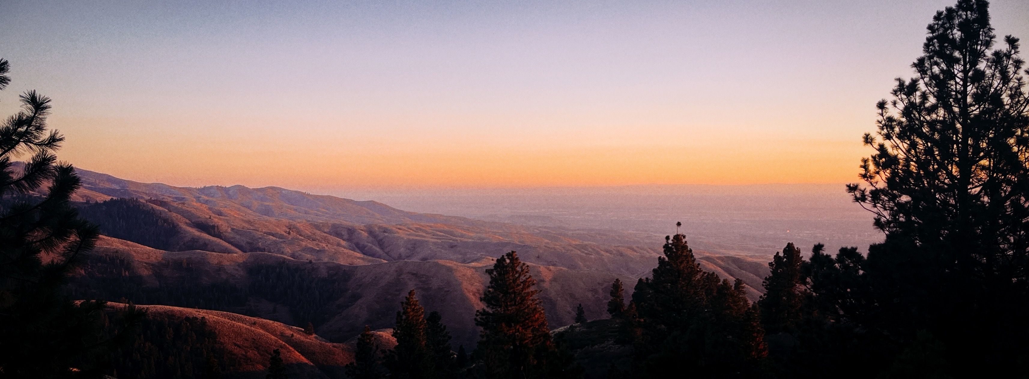 A wide-angle photograph taken from a mountain above Boise, Idaho. Hills stretch out in the distance. They look purple because of the sunset, which glows orange on the horizon.