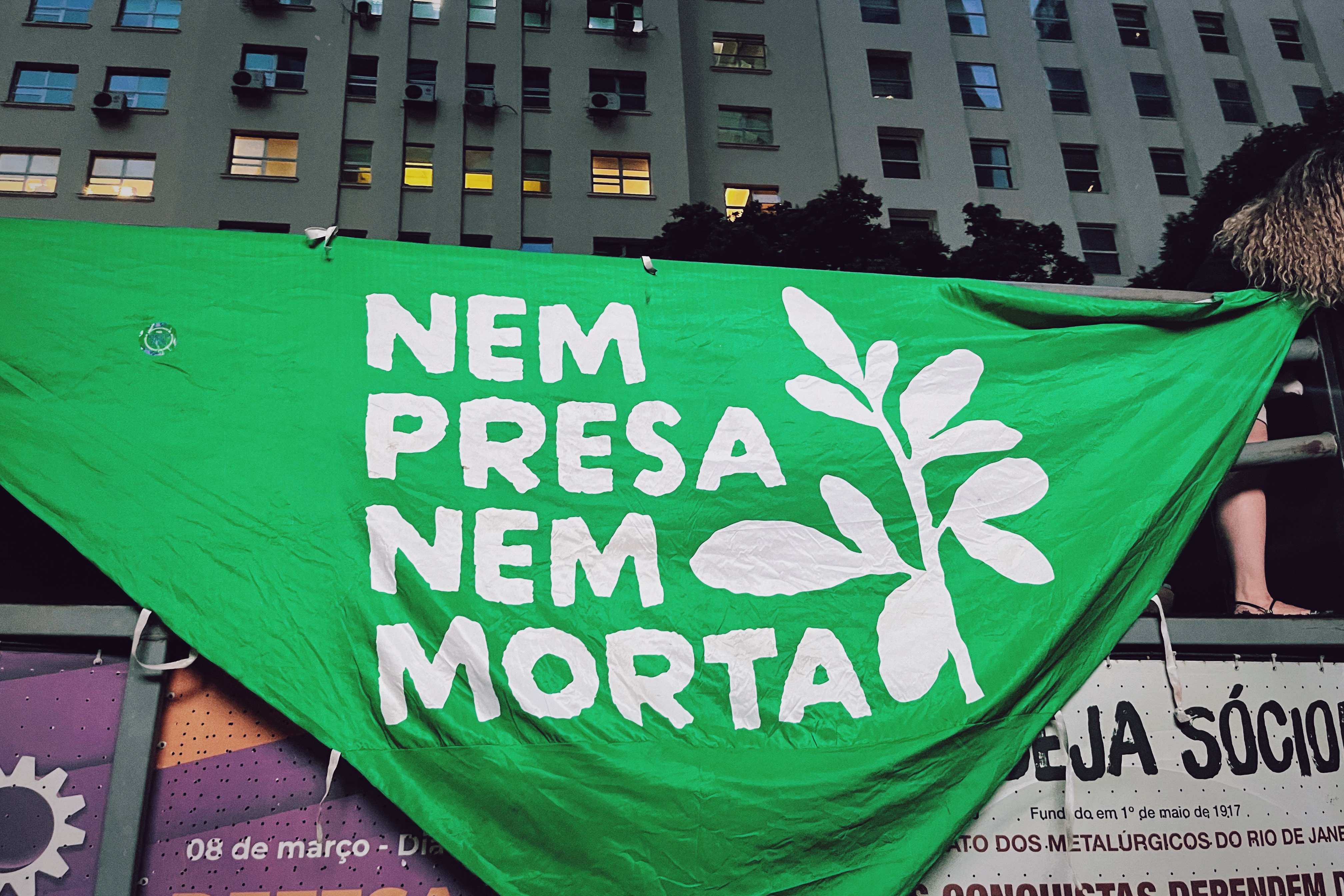 Photograph of a large green banner that says NEM PRESA NEM MORTA, hanging from the side of a bus during the Women's Day march in Rio de Janeiro.