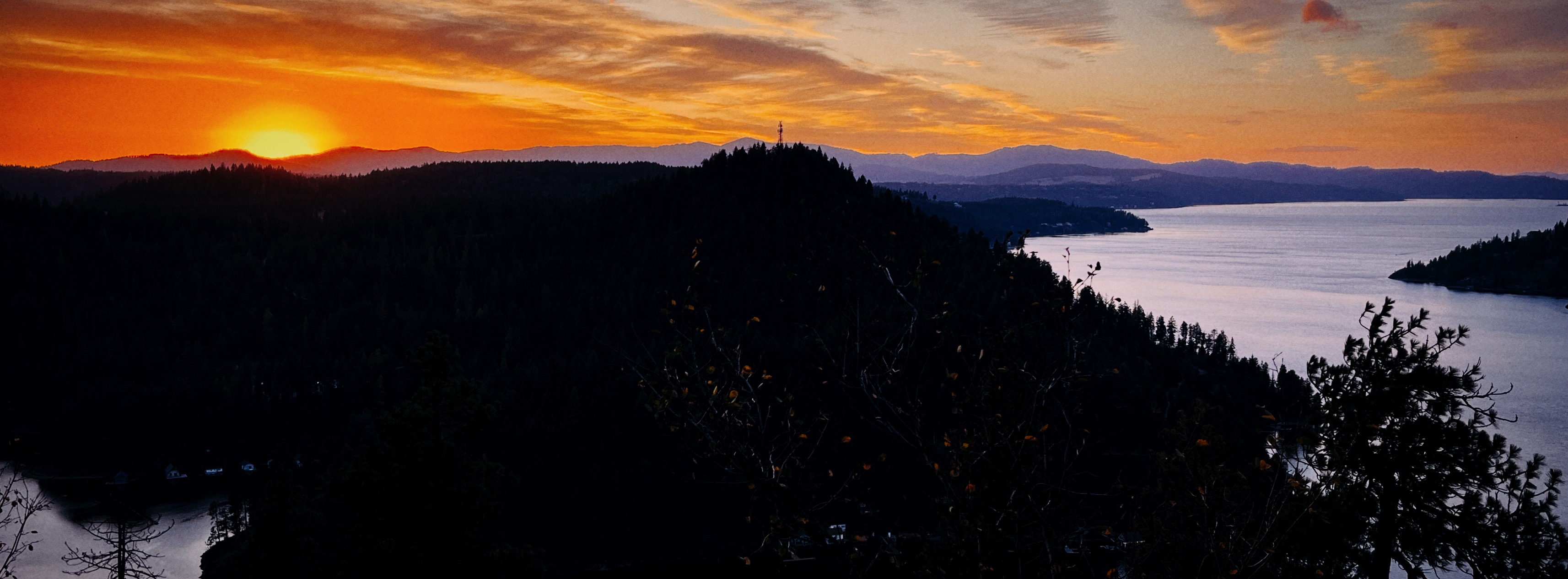 A wide-angle photograph of sunset over Lake Coeur d'Alene. The sun glows yellow just above the horizon, wiht notes of red, orange, yellow, and pink in the sky. The foreground of the photo shows trees, which are so dark they look black, and the winding blue waters of the lake. There are blue-purple mountains in the distance.