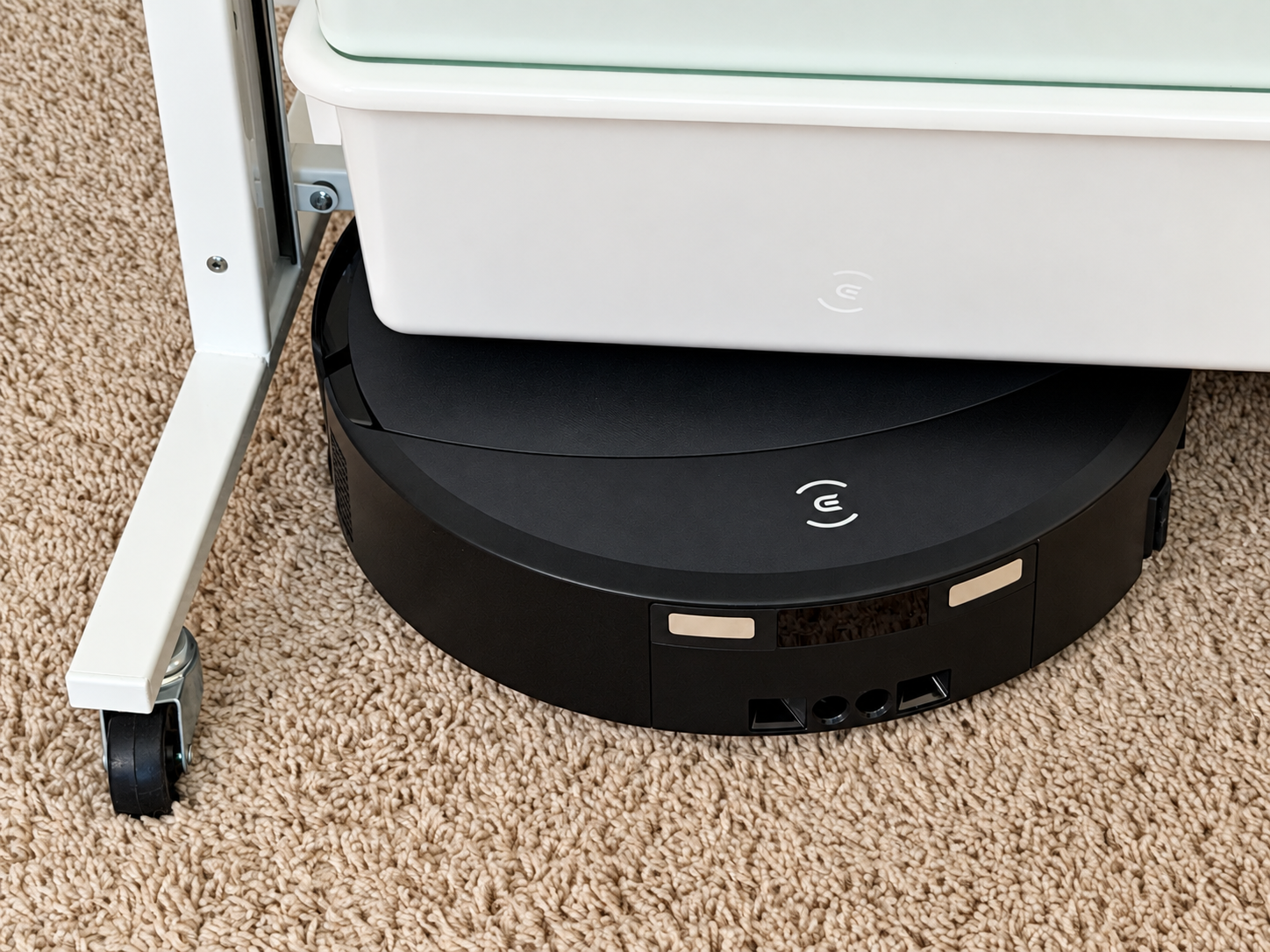 Ecovacs Deebot T90 Pro Omni cleaning beneath a low white piece of furniture on beige carpet.