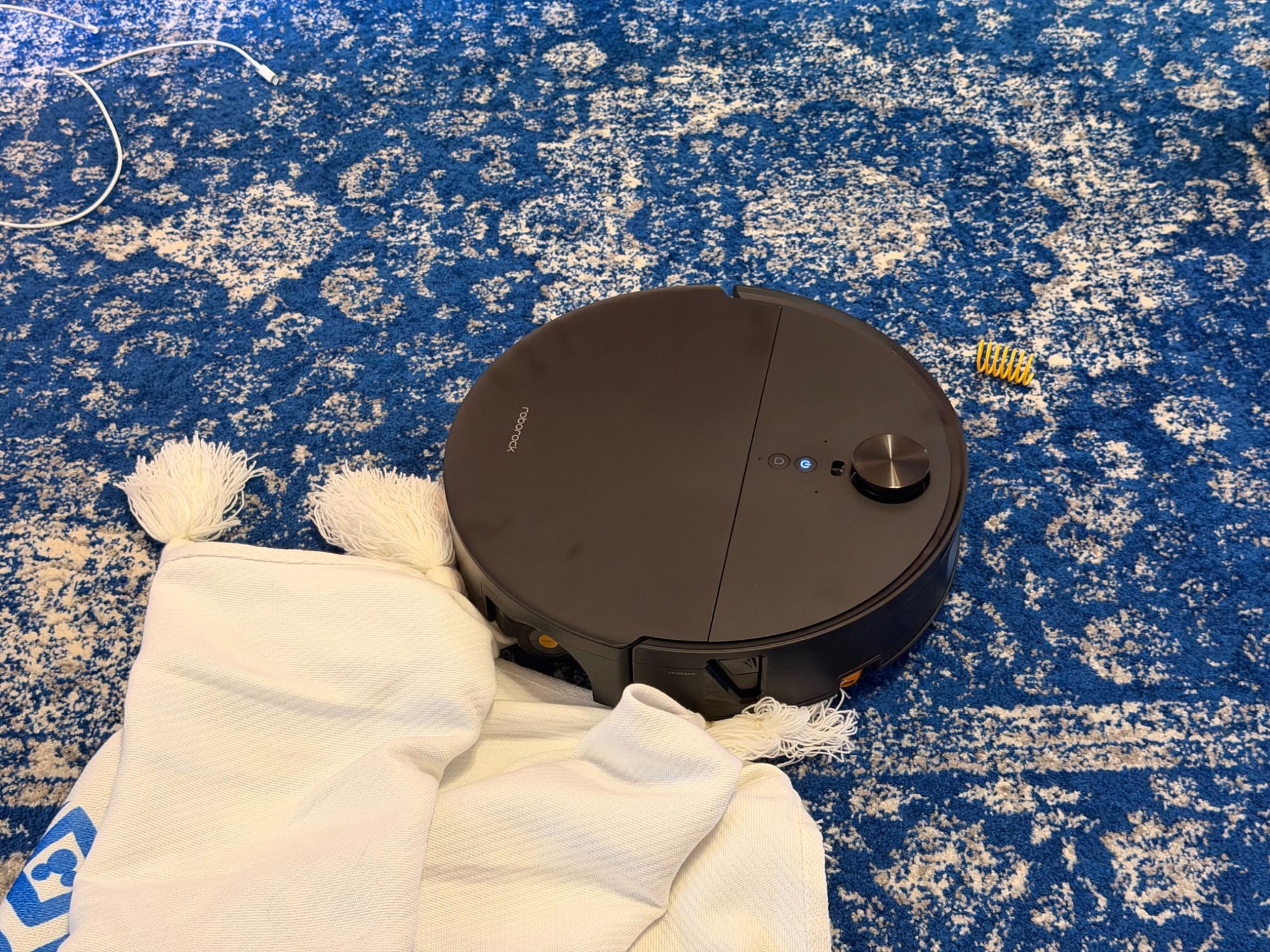 Roborock Saros 20 Flow robot vacuum caught in the tassels of a white throw blanket on blue patterned carpet, with a charging cable and yellow cat spring nearby.
