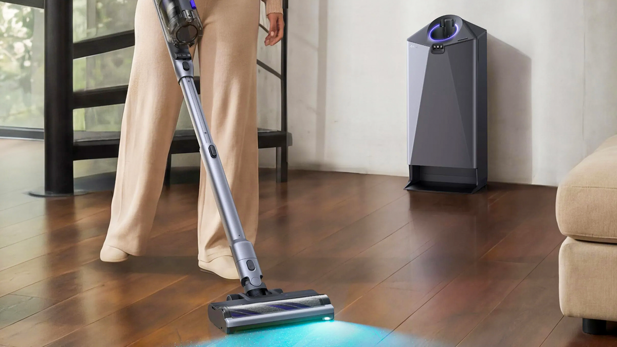 A person uses a Narwal V50 Cube cordless vacuum on a wood floor, with an aqua light illuminating dust and the self-emptying dock nearby.