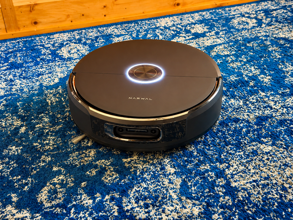 A black Narwal Flow 2 robot vacuum sits on a blue-and-white patterned area rug near a wood floor, with its illuminated control ring visible on top.