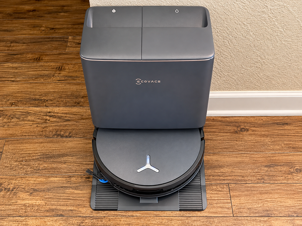 Ecovacs Deebot T90 Pro Omni robot vacuum docked at its dark gray all-in-one base station on a wood floor.