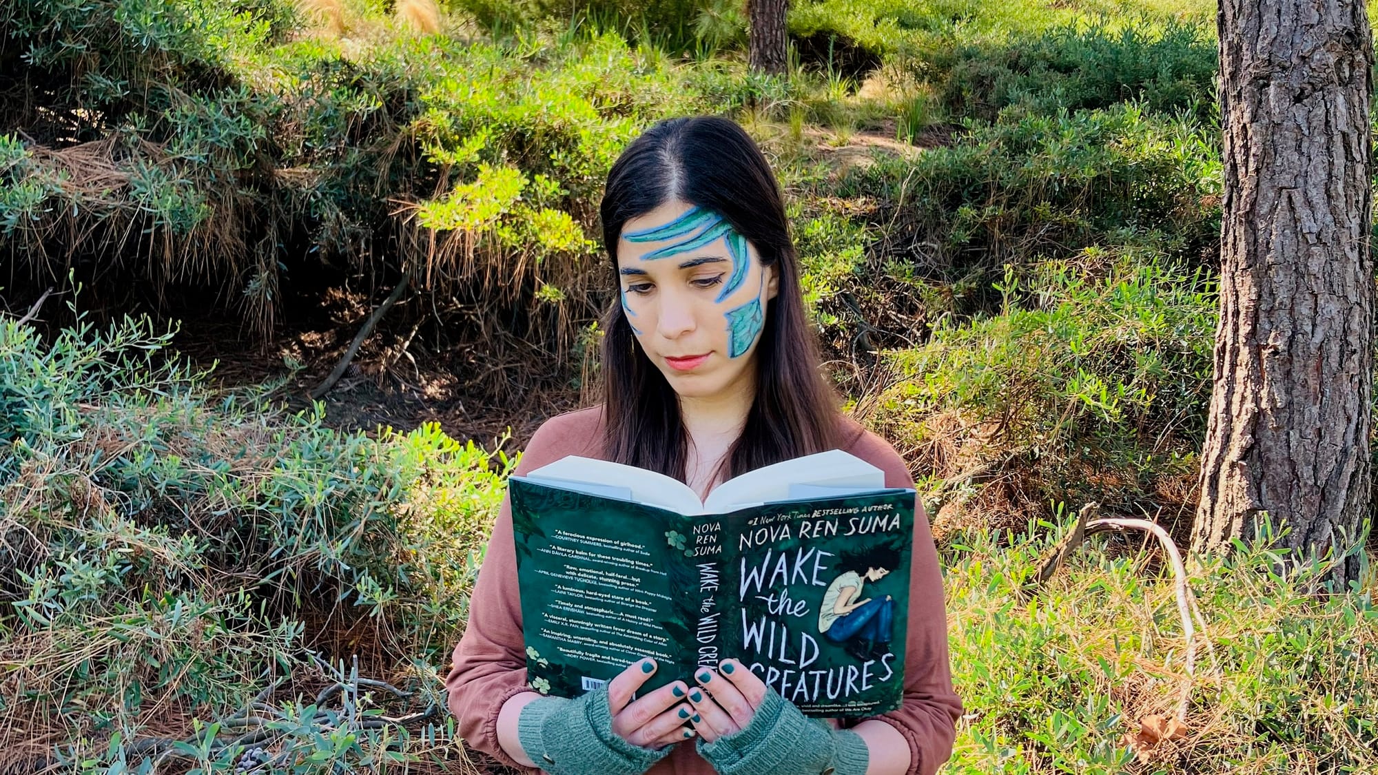 A-M reading Nova Ren Suma’s novel WAKE THE WILD CREATURES. They are standing in front of trees and undergrowth and have blue and green vines painted on their face.