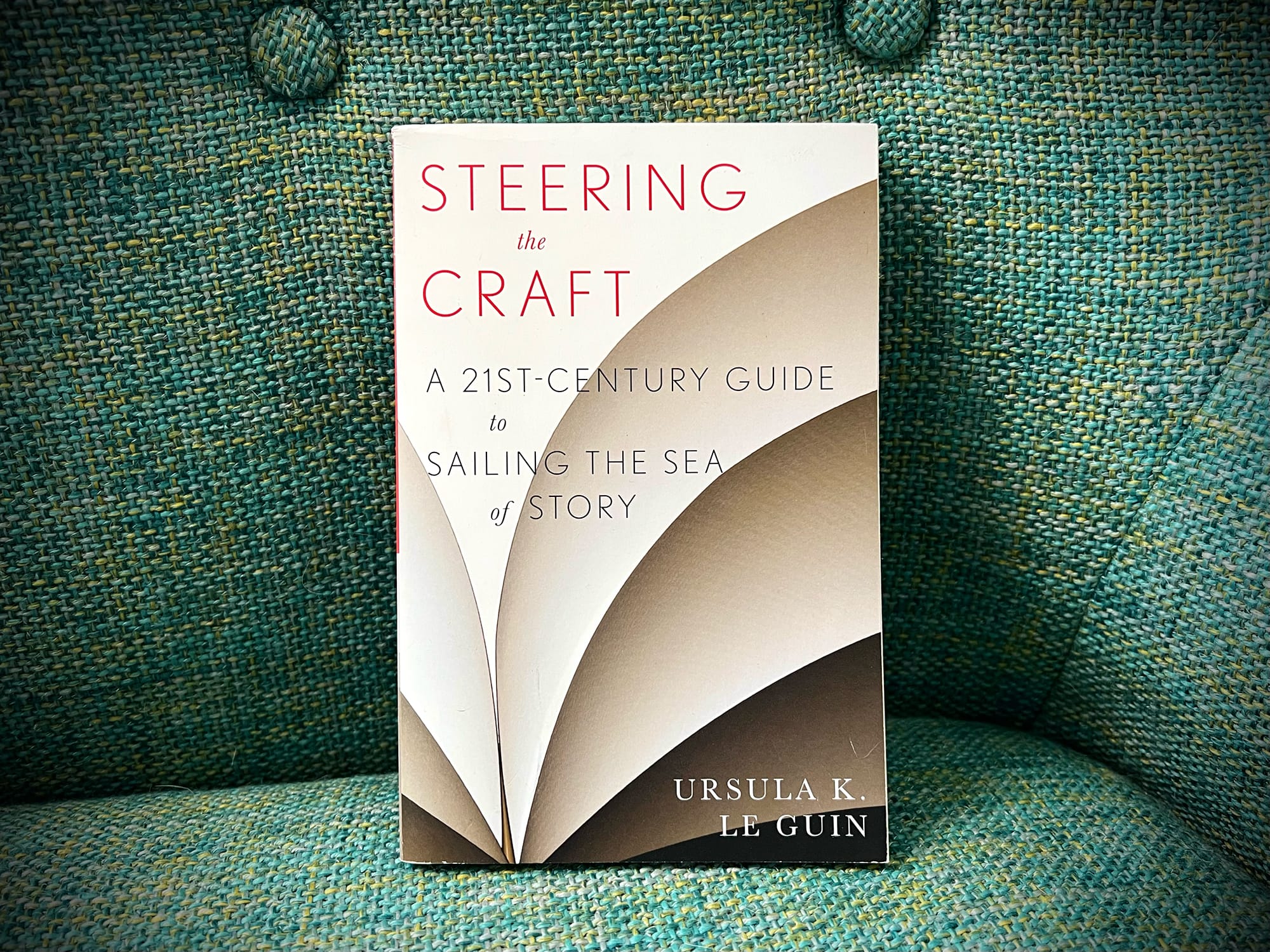 A photograph of the book Steering the Craft: A 21st-Century Guide to Sailing the Sea of Story by Ursula K. Le Guin. The book is upright against the back of a green upholstered chair.