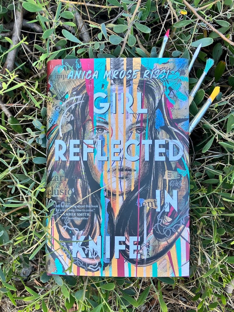 Hardcover of Anica Mrose Rissi's GIRL REFLECTED IN KNIFE, resting on top of plants, with paint brushes sticking out of the pages. The brushes have red, white, teal, and gold paint on them, matching the colors of the cover.