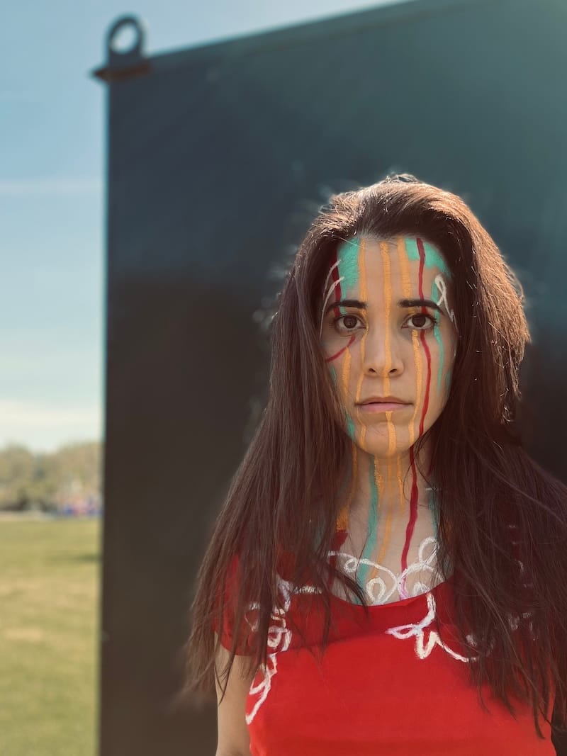 A-M stands in front of a green electrical box, with a field and trees in the background. Their face is painted with swirls, squiggles, patches, and lines of aqua, red, gold, and white. A red shirt painted with white squiggles is visible in the frame.