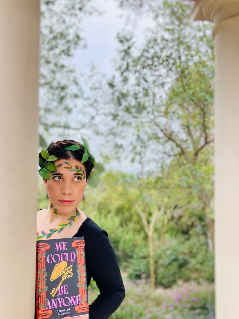 Author leaning against white stone column holding book with vining leaves painted on face and leaves woven into braided hair.