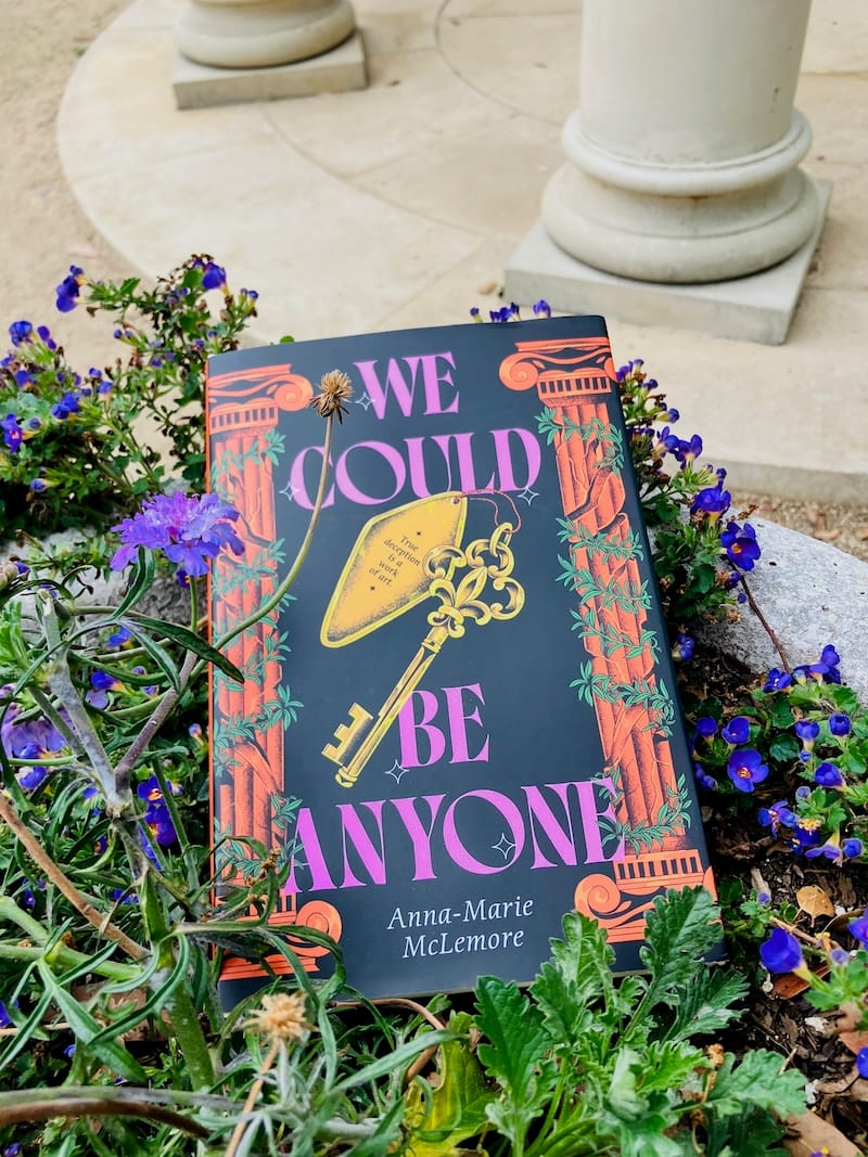 Hardcover of Anna-Marie McLemore’s We Could Be Anyone among vining plants with purple flowers with columns visible in background.