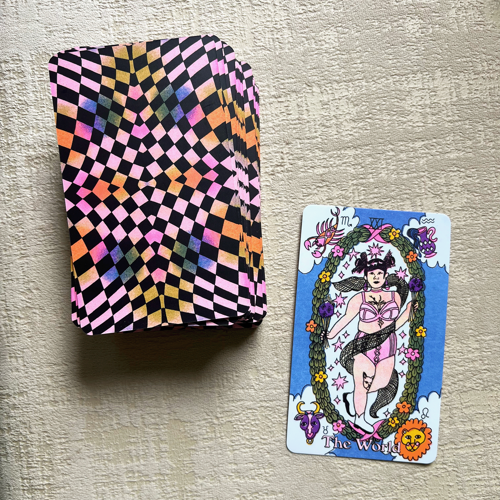On the left is a facedown deck of Tarot cards with a trippy checkered pattern on the backs. On the right is The World card, which depicts a femme-presenting figure holding bouquets and standing inside a giant wreath. In the four corners of the card are a lobster, a jug of water, an ox, and a lion.