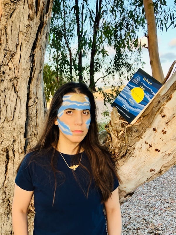 Author with moon and cloud pattern painted on face, standing near WHEN THE MOON WAS OURS paperback resting on tree branch