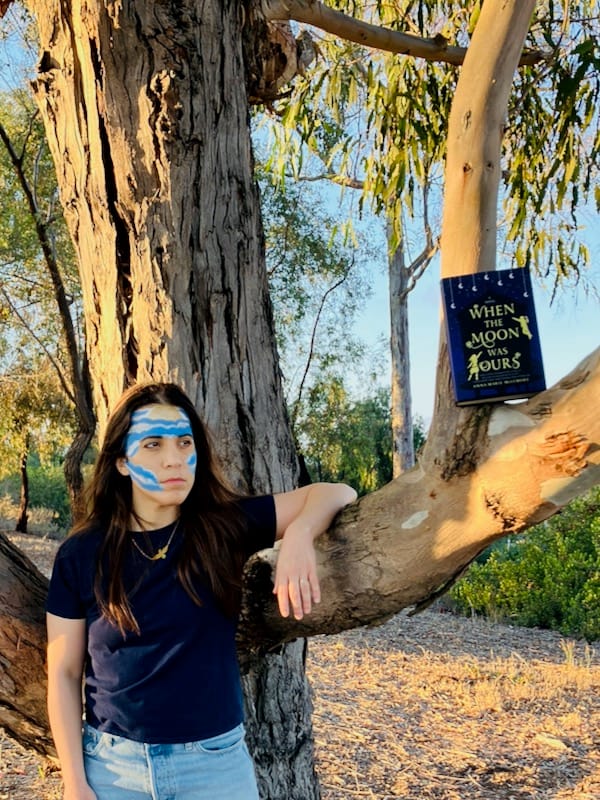 Author with moon and cloud pattern painted on face, leaning against tree near WHEN THE MOON WAS OURS hardcover resting on branch