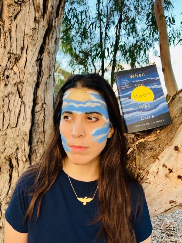Close up of author with moon and cloud pattern painted on face, standing near WHEN THE MOON WAS OURS paperback resting on tree branch
