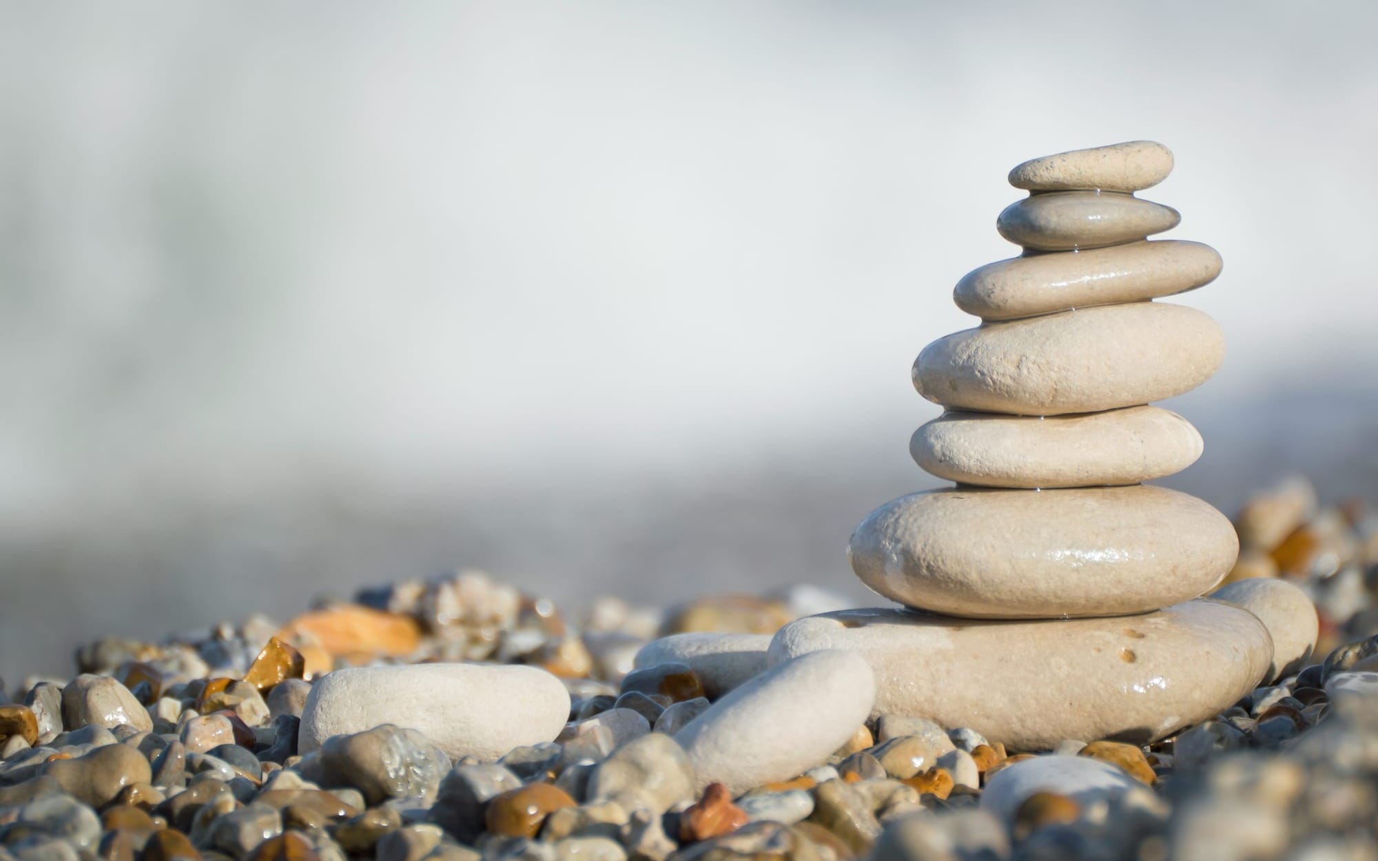 A close-up photograph of several smooth, light-colored stones perfectly stacked in a balanced tower on a pebble beach, representing zen, inner harmony, and alignment.