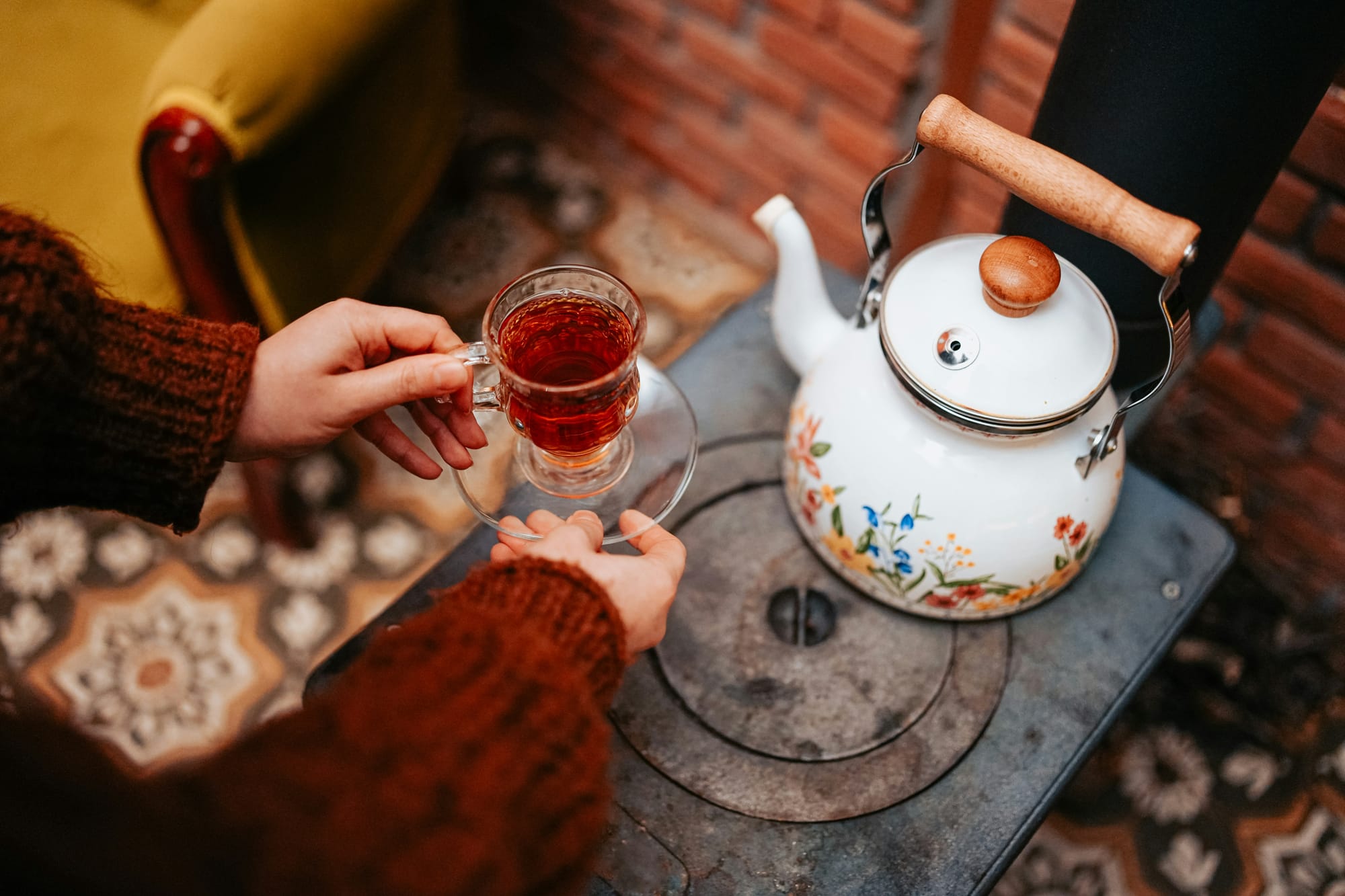 A warm, rustic overhead photograph showing two sets of hands wearing cozy knit sweaters gently sharing a clear glass cup of hot amber tea next to a vintage floral teapot.