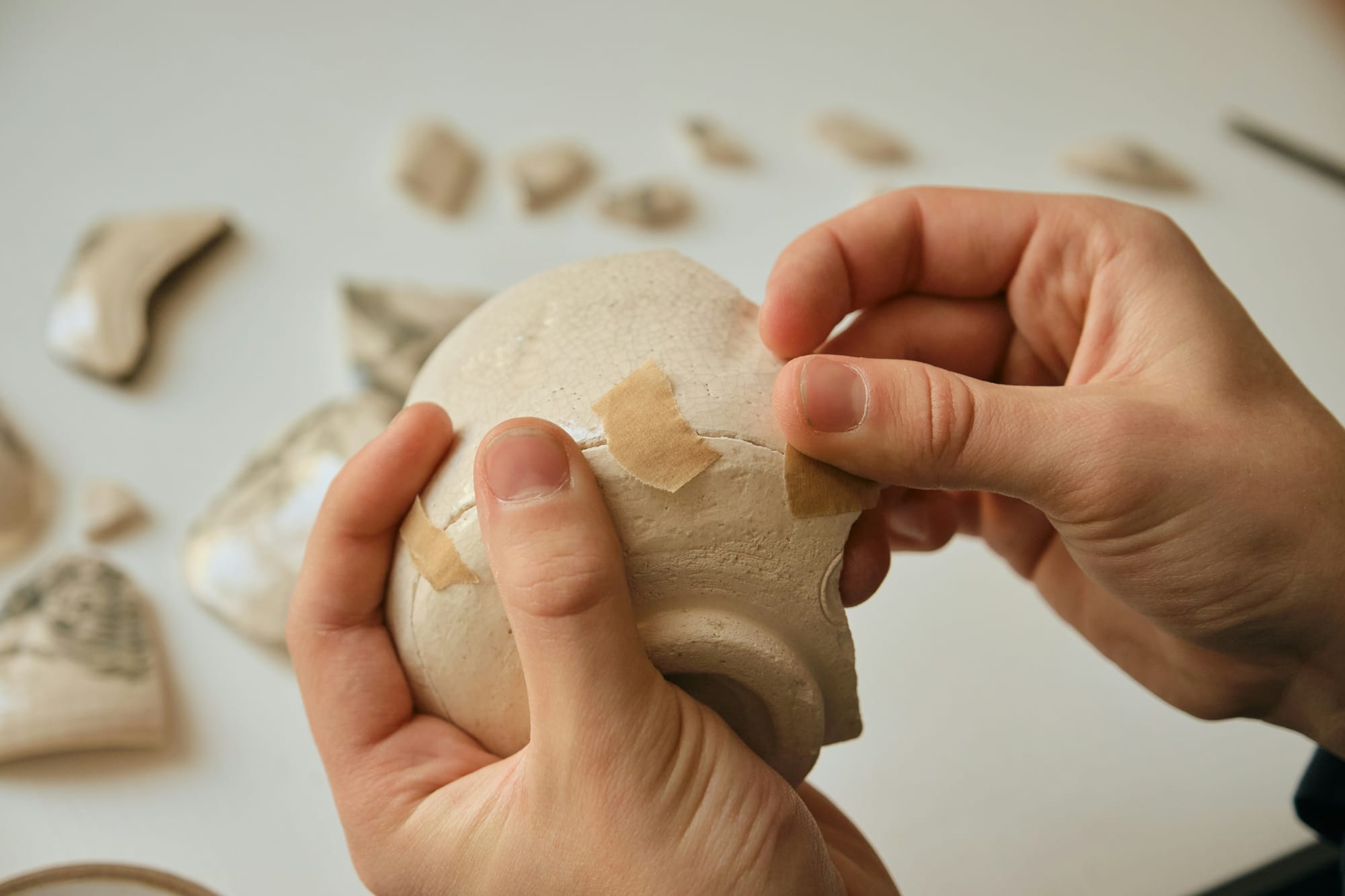A close-up of hands applying paper tape to a broken ceramic bowl, representing makeshift emotional coping mechanisms and the hidden pain of betrayal.