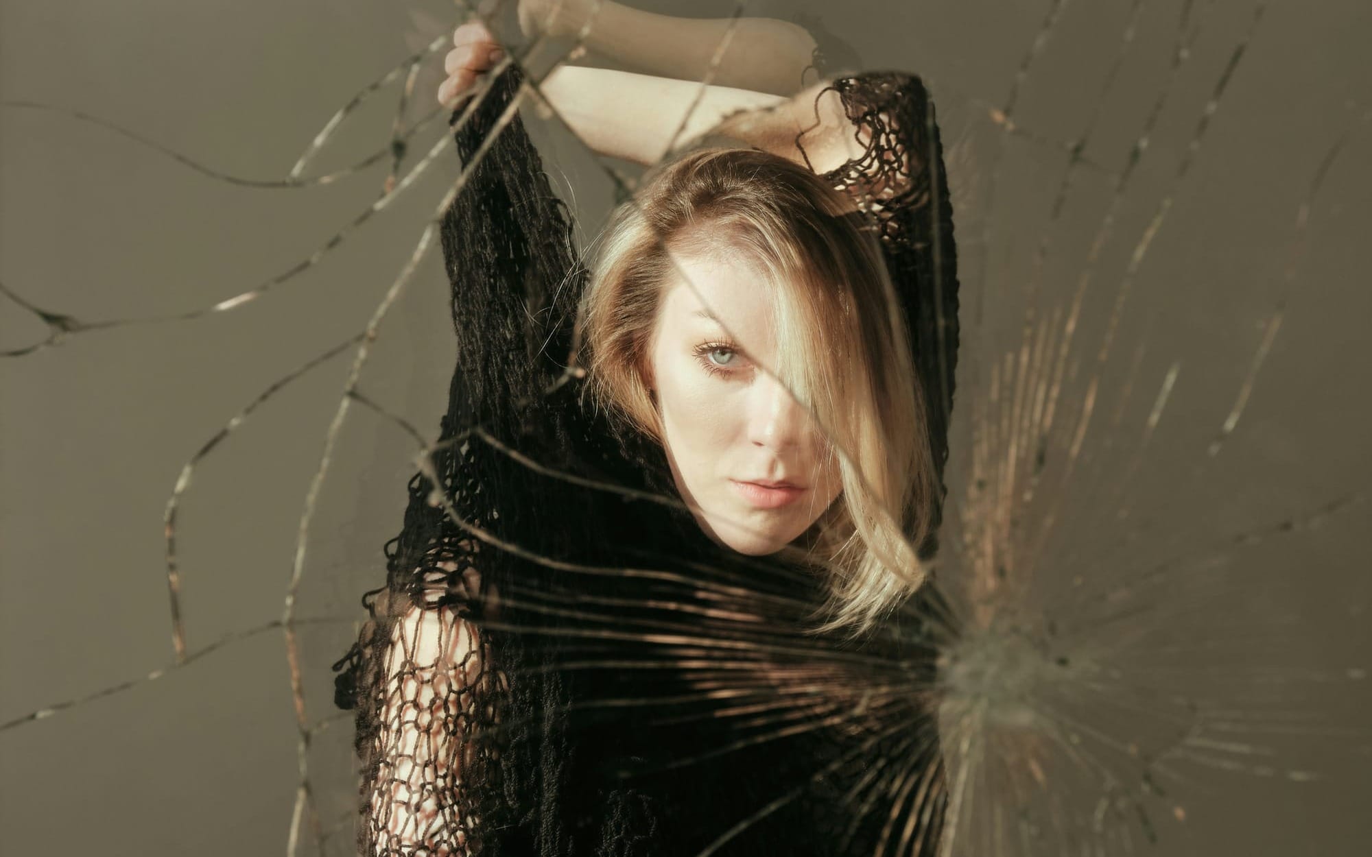 A dramatic conceptual portrait of a woman looking directly at the camera through a shattered pane of glass, with cracks splitting her reflection into fragments, symbolizing the fragmented self and identity loss.