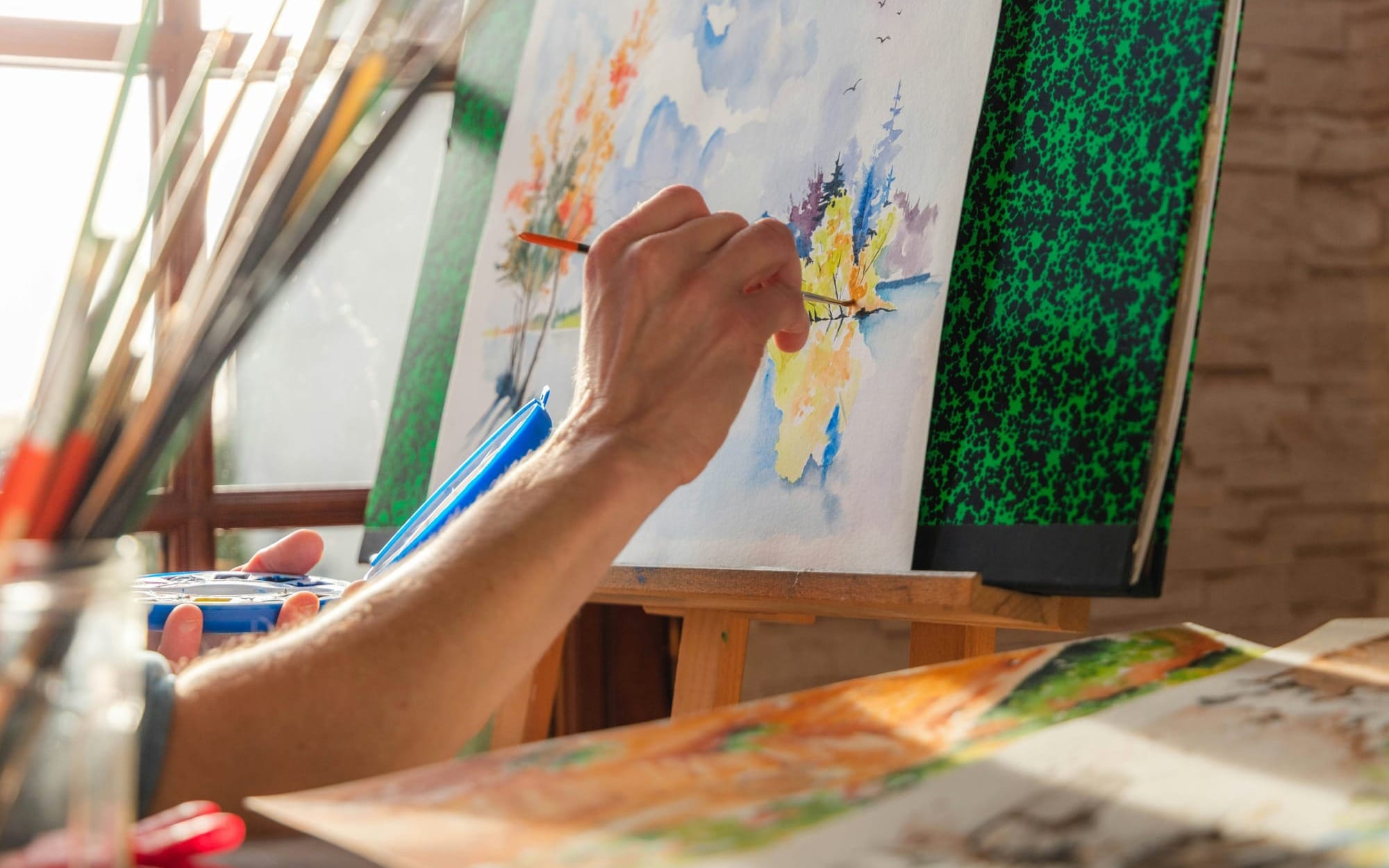 A close-up of an artist's hand using a paintbrush to paint a colorful landscape watercolor on an easel.
