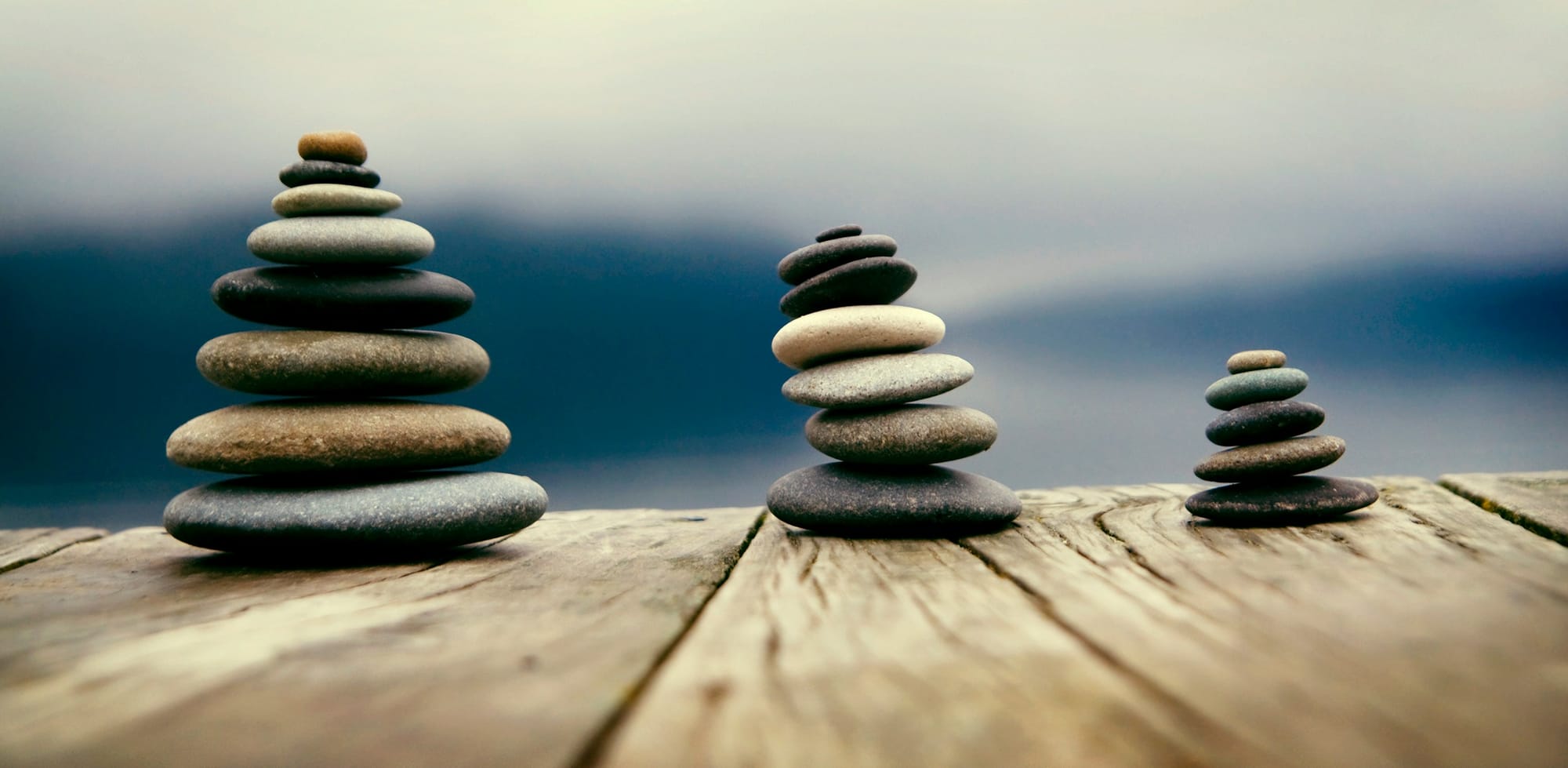 Three balanced stacks of smooth, weathered zen river stones arranged on a rustic wooden pier against a soft, misty blue water background, representing structural stability.