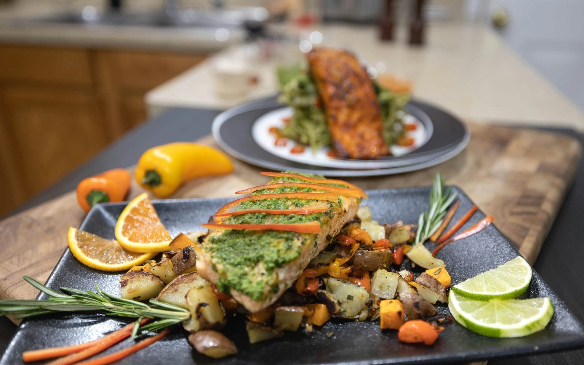 A plated salmon fillet topped with green herb pesto, served over a bed of roasted potatoes and fresh peppers.