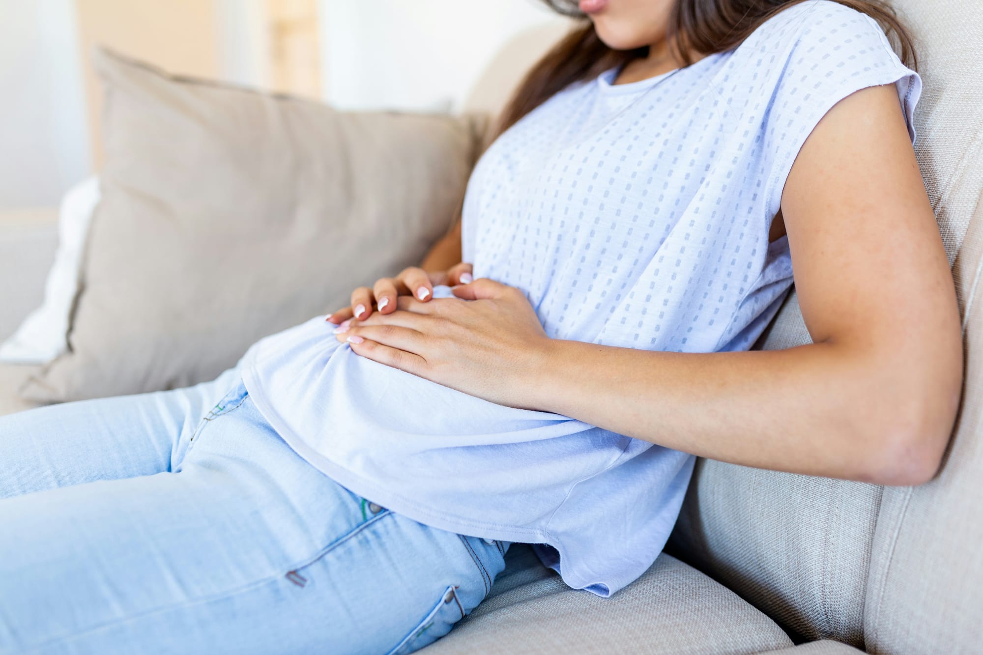 Woman resting on a couch while holding her lower abdomen, representing chronic UTI discomfort and persistent pelvic symptoms.