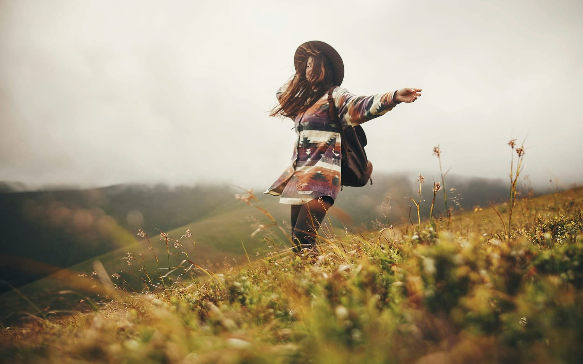 A joyful woman with a wide-brimmed hat and backpack walking on a windy mountain ridge with her arms outstretched, embodying self-affirmation, emotional freedom, and protecting her self-integrity.
