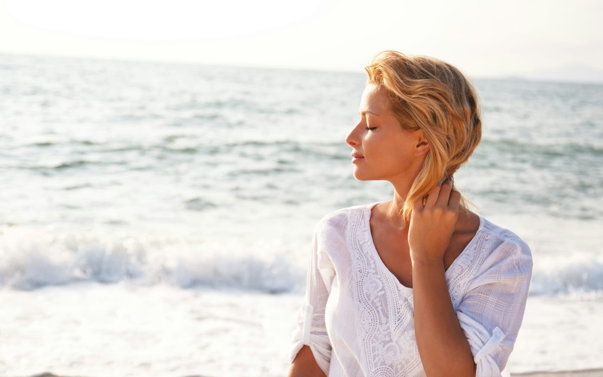 A serene profile photograph of a woman with closed eyes standing by the ocean in warm, bright sunlight, breathing in the sea breeze and experiencing deep mental clarity, peace, and connection with her inner voice.