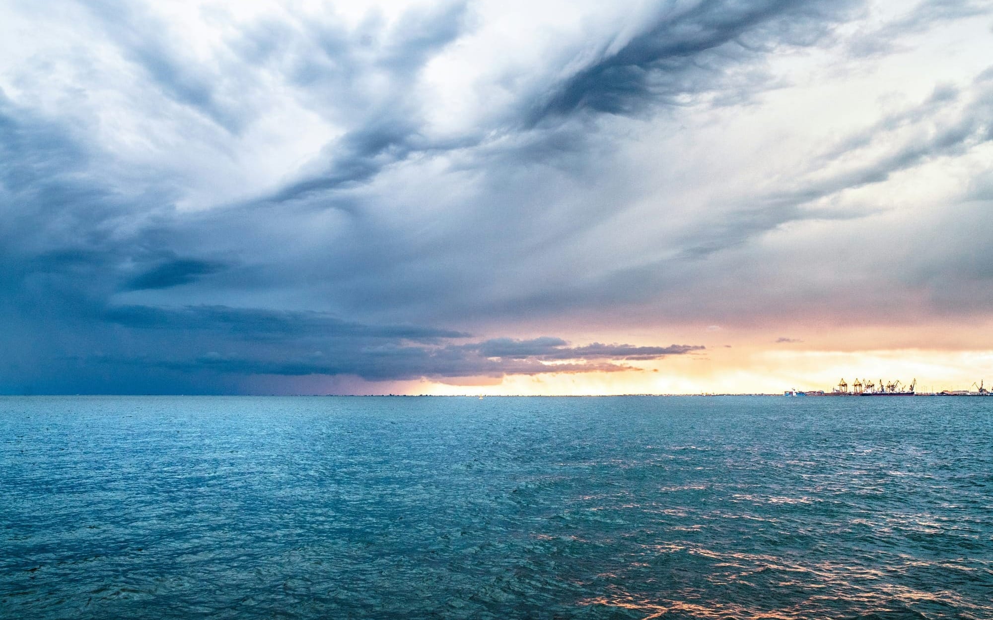 A dramatic seascape featuring vast blue ocean water under a heavy, turbulent storm cloud formation on the left, contrasted by a beautiful, bright golden horizon breaking through on the right, symbolizing overcoming the internal storm of imposter syndrome.
