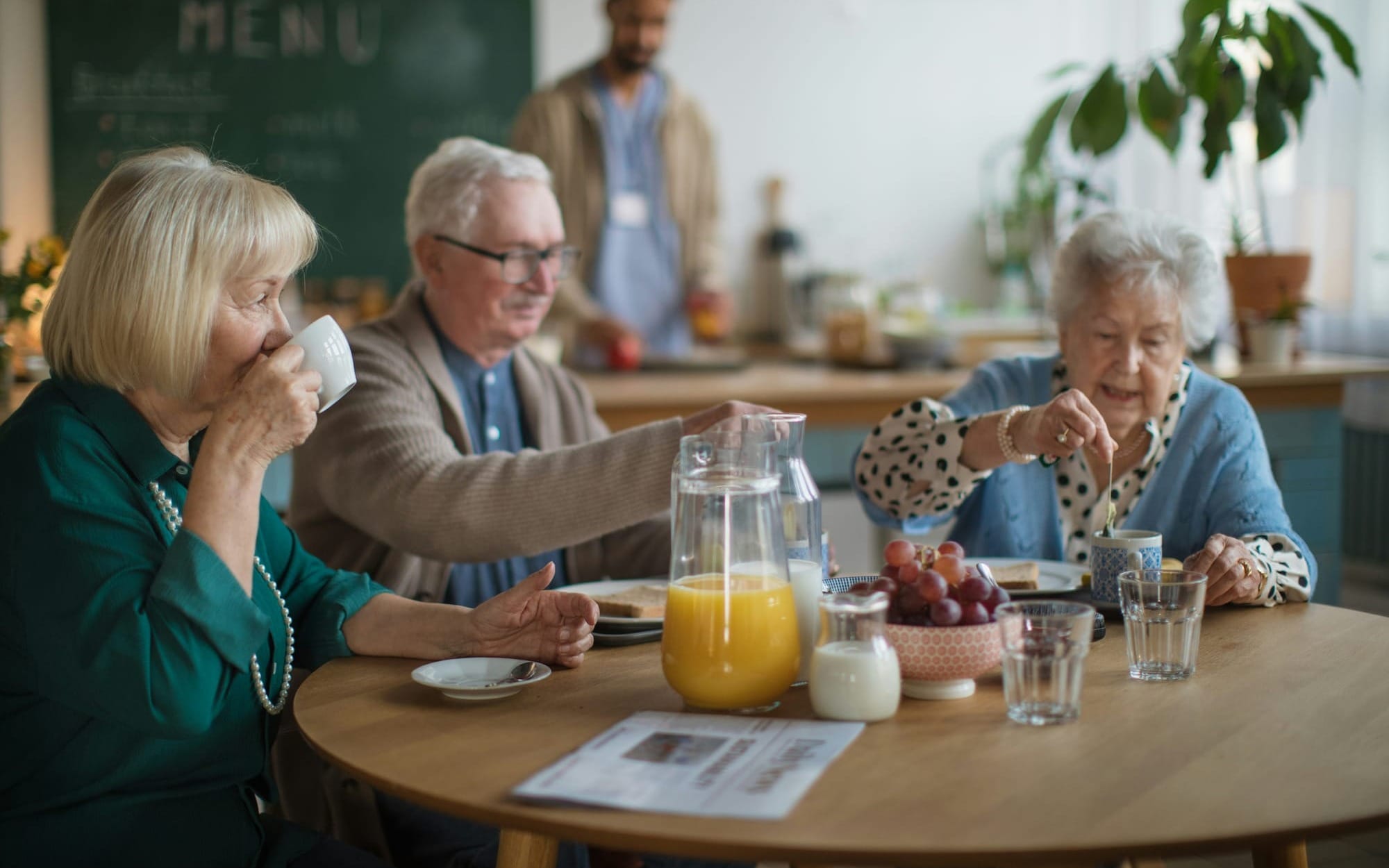 Three older adults sharing a calm breakfast in a bright community kitchen, surrounded by fresh juice, fruit, and gentle conversation — a visual of social connection and nourishment central to Blue Zones living.
