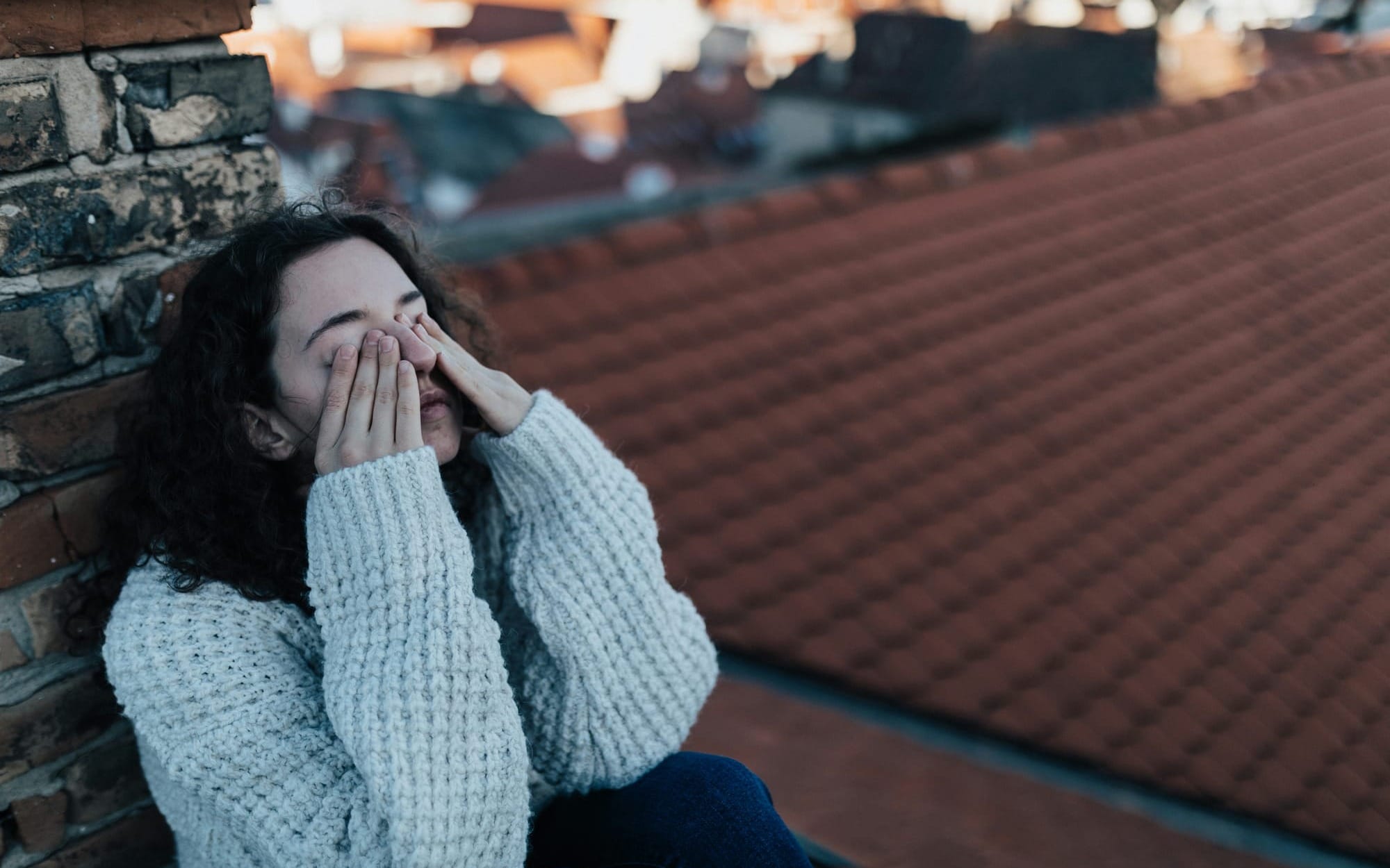 A young woman sitting outside on a rooftop with her back against a brick wall, covering her face with her hands in a moment of exhaustion, capturing the feeling of status anxiety and burnout.