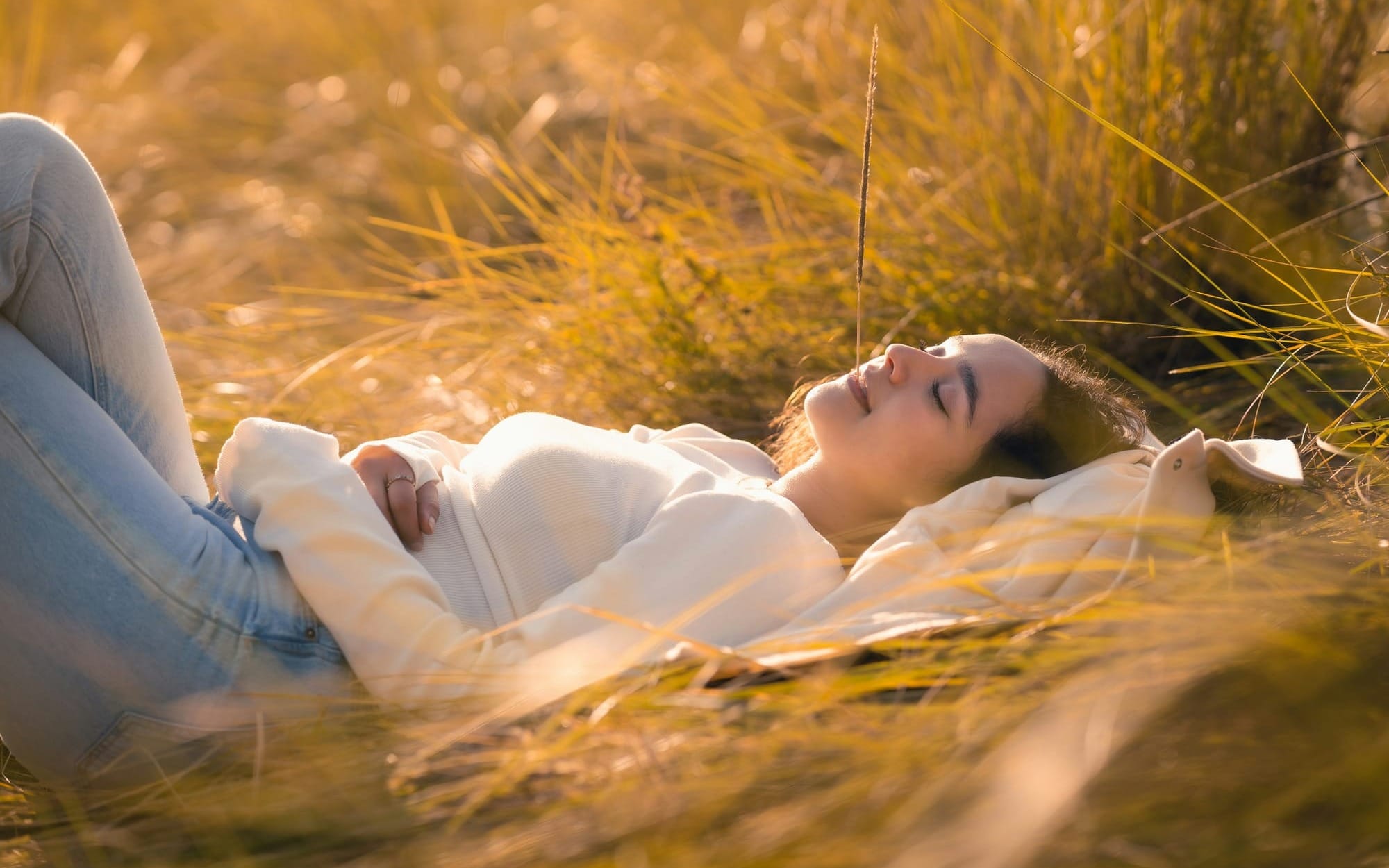 A young woman lying down peacefully in a field of tall golden grass with her eyes closed and a soft smile, bathed in warm sunset light, capturing the essence of self-acceptance and the true self.