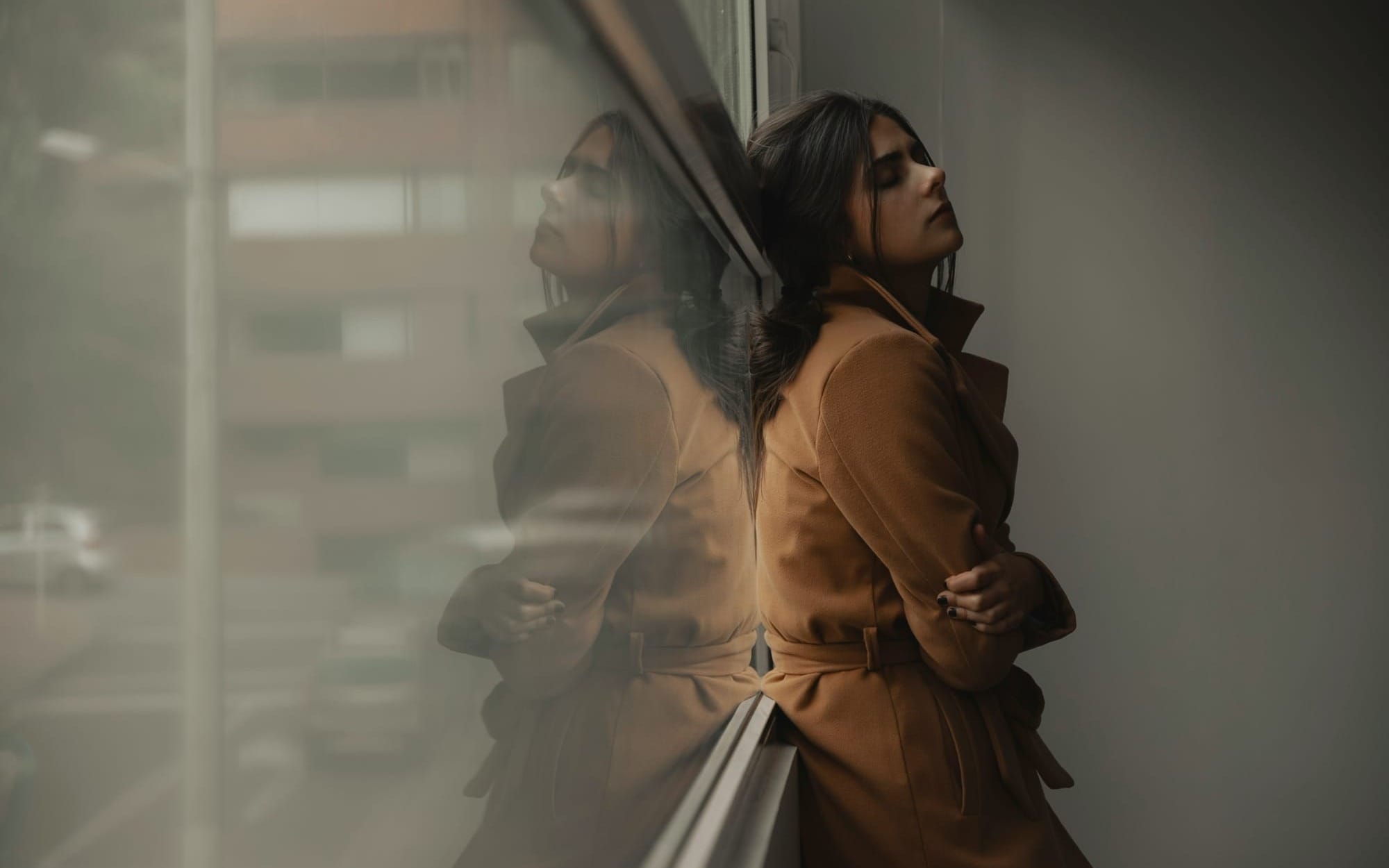 A woman in a brown coat leaning against a large window with her arms crossed and eyes closed, showing her clear profile next to a soft, moody reflection of herself in the glass, symbolizing the imposter syndrome and split self-perception.